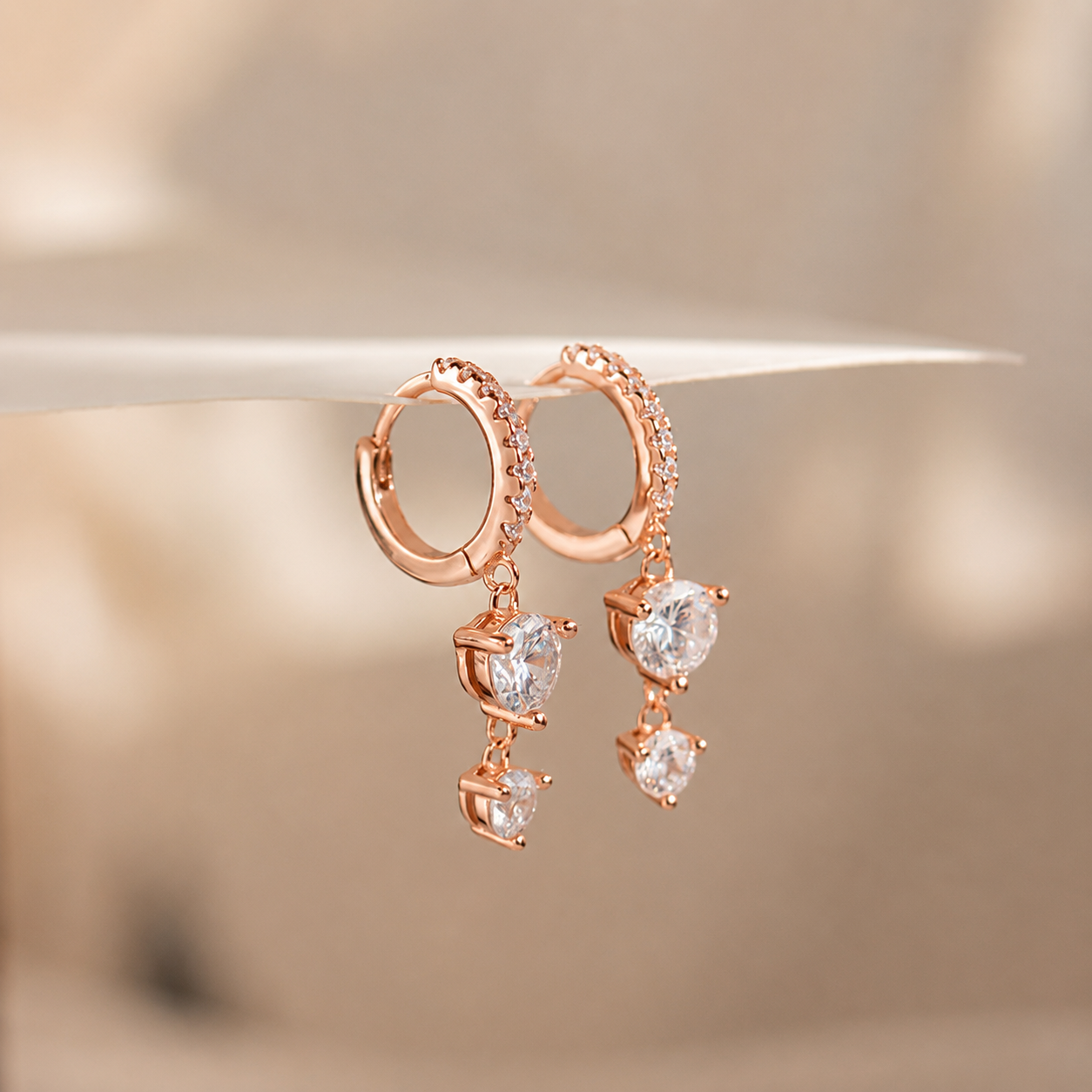 Rose gold huggie hoop earrings with small clear stones along the outer edge and dangling clear gemstone charms, shown against a soft beige background.
